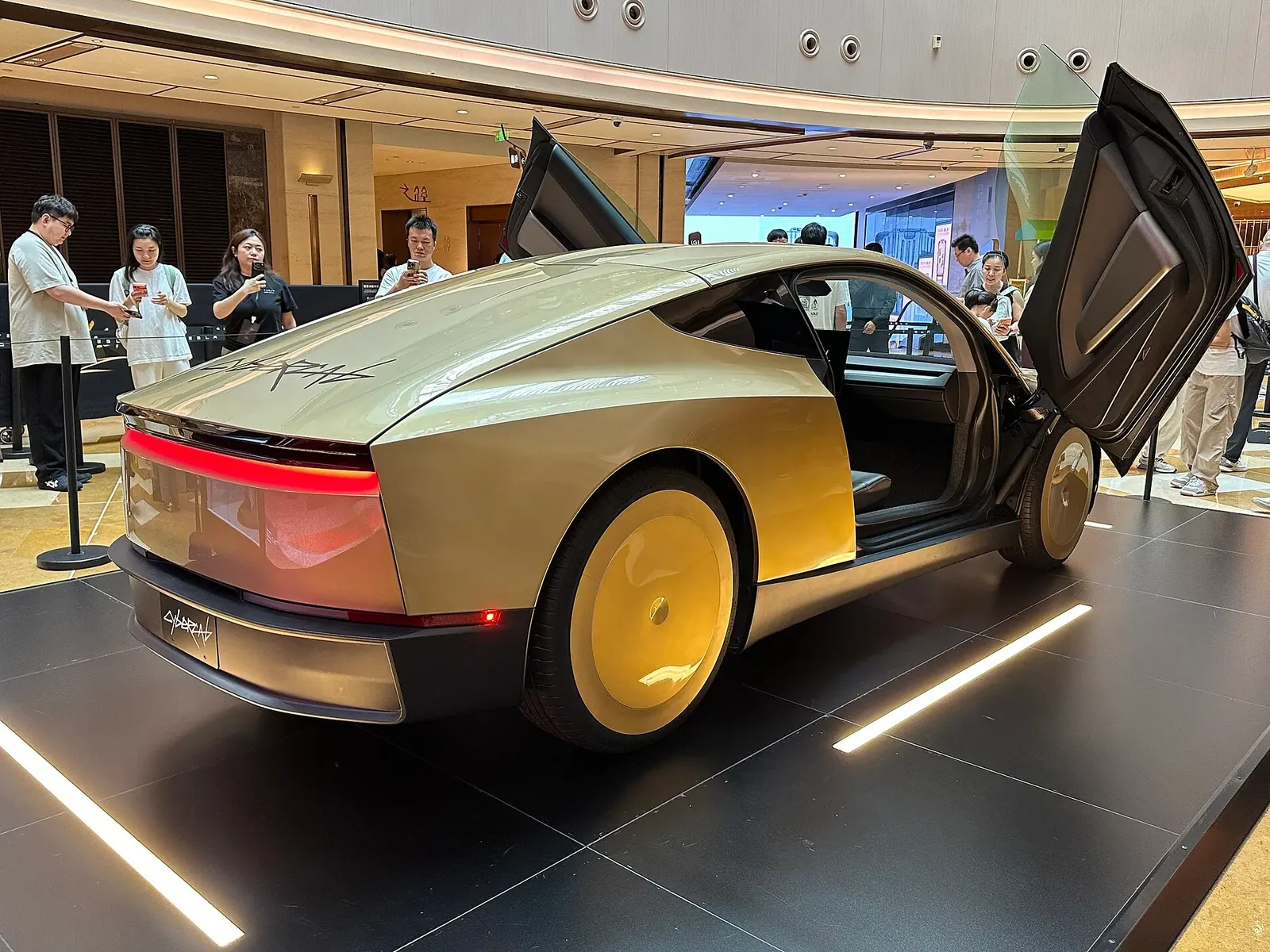 Gambar: JustAnotherCarDesigner / Wikimedia Commons (CC0) Bahagian belakang Tesla Cybercab warna emas dengan lampu belakang merah sebaris, pintu butterfly terbuka, dipamerkan dalam mall