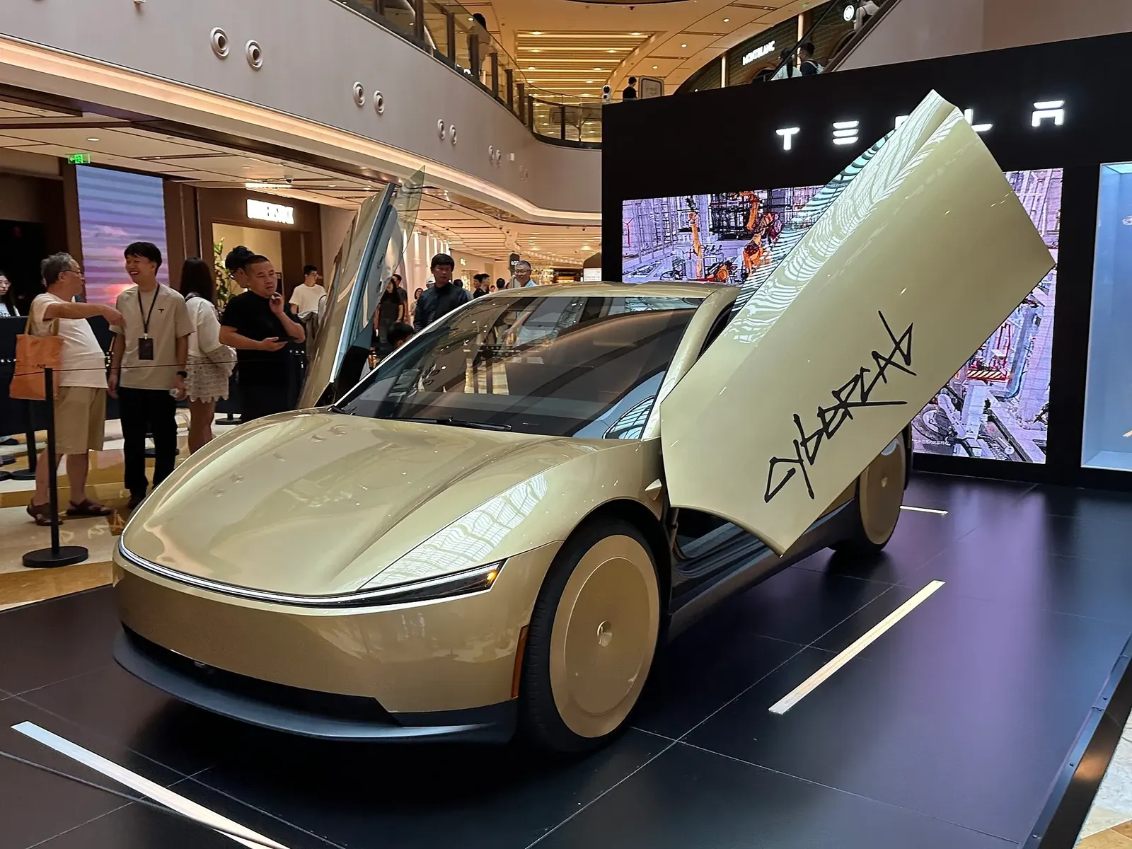 Gambar: JustAnotherCarDesigner / Wikimedia Commons (CC0) Tesla Cybercab warna emas dengan pintu butterfly terbuka dipamerkan atas pentas di sebuah mall dengan pengunjung di sekeliling