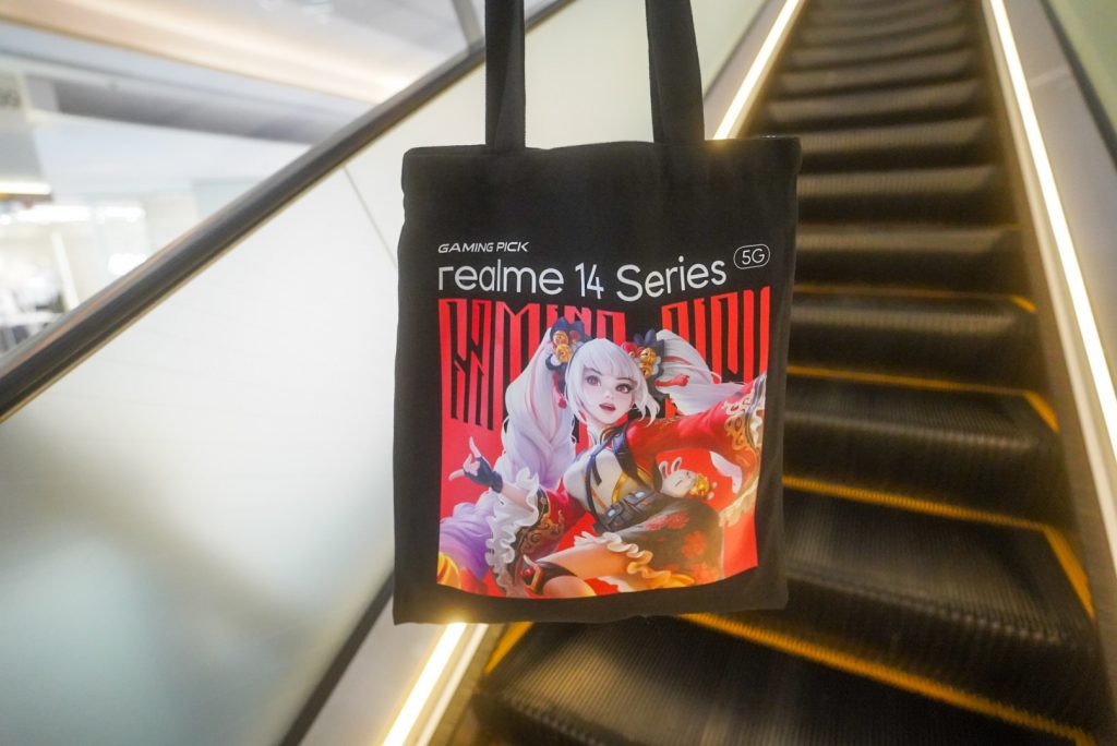 realme 14 Series Gaming Pick tote bag hanging in front of a mall escalator
